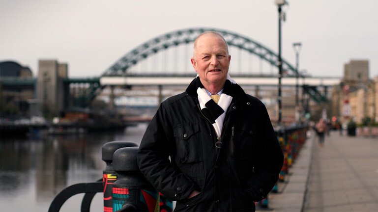Sir Doug Turnbull standing by the river in Newcastle with the Tyne bridge in the background