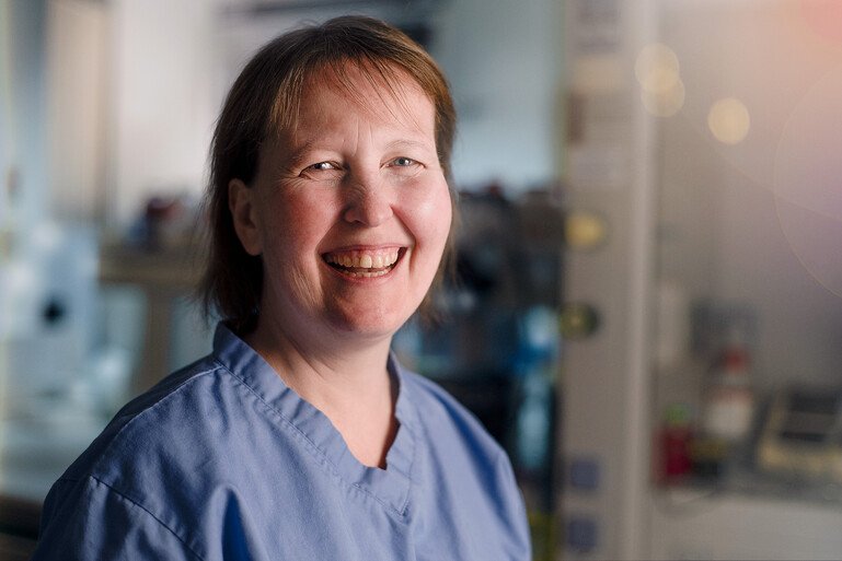 Louise Hyslop wearing blue lab scrubs in a clinical research setting, with laboratory equipment blurred in the background.