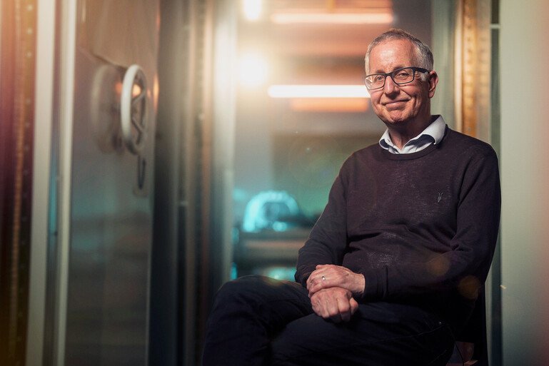 A seated portrait of Gareth Barnes in a corridor, wearing glasses and a dark jumper, with soft lighting in the background.