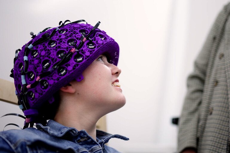 Liberty wears a purple sensor-equipped OPM-MEG helmet while seated indoors and looking upward during a brain scanning session