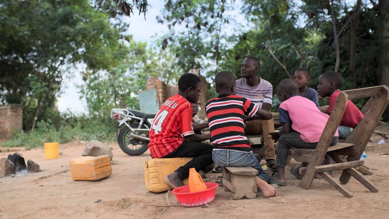 An adult and several children sit together on wooden seats outdoors.