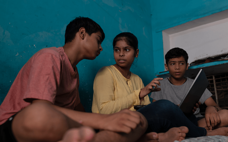 Three children sit on a bed and look at a laptop