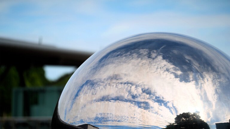 Clouds, trees and buildings reflected in a curved, mirrored surface outdoors.