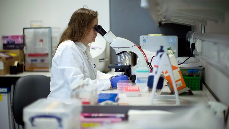 A person in a white laboratory coat and blue gloves uses a microscope at a laboratory bench.