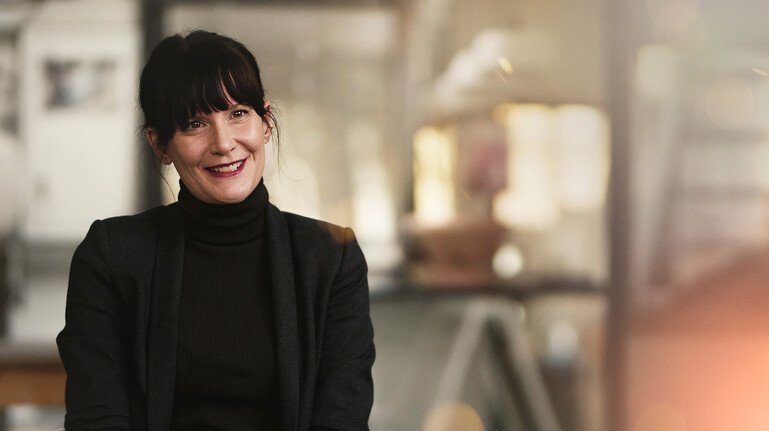 Amy Hardy sits indoors in a black jacket and high-neck top, with a softly blurred background.