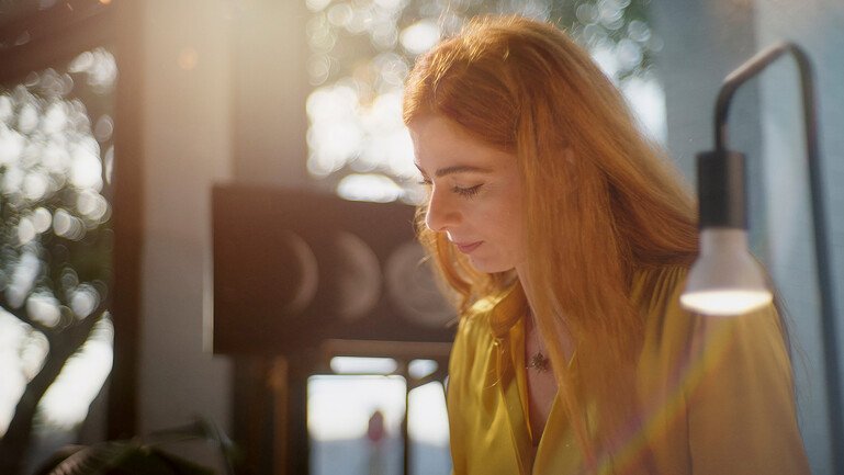 Anna Wojdecka looks down beside a lit desk lamp, with sunlight visible behind her.