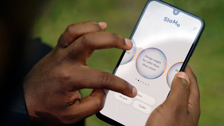 Hands hold a phone showing SloMo thought bubbles, including one that reads “things might be safer than they seem”.