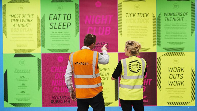 Two people facing wall full of posters with information about sleep and health, as part of the workshop and installation Night Club.