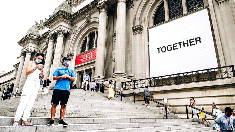People wearing protective masks stand on the steps of the Metropolitan Museum of Art in New York which displays a banner with the word 'together'.