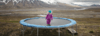 A girl stands on a trampoline in the middle of a mountainous landscape.