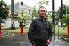 Sudarmono Djoko Nugroho stands just right of centre. We see him from the waist up. He wears a dark coloured shirt and stands in front of palm leaves on a paved road.