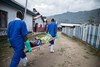 Two medical workers in bright blue uniforms carry a patient on a stretcher down a gravel path in a town in the Democratic Republic of the Congo.