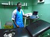 Edrisa Sinjanka, a midwife in The Gambia, stands beside a bed inside a maternity clinic with his hand on a monitor.