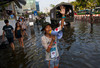 A person wearing the Red Cross badge on their arm stands on a flooded road, talking through a megaphone. Soldiers and civilians along with a military vehicle can be seen in the background.  