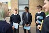 Five secondary school pupils wearing school uniforms are gathered in a circle smiling and chatting whilst holding books.