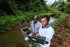 A man and a woman stand beside a river in Kenya, they're holding scientific sampling equipment and testing samples from the river and soil.