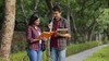 Two students talking to each other and looking at a book. They are stood outdoors on a path lined by trees.