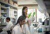 A woman in a white lab coat and bright green gloves hold a petri dish up to the light to examine it. She's standing in a busy lab surrounded by her peers.