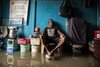 A man sits in his room with his feet up on a paint bucket to avoid floodwater.